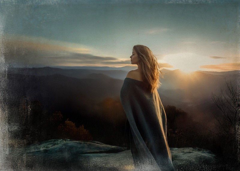A mysterious witch stands atop the Smoky Mountains at sunset in late fall, her dark wool cloak billowing in the wind as she gazes into the distance, framed by golden light and autumnal hues.