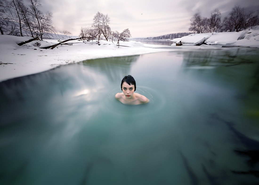 A pale young woman with wet black hair swims in an icy blue-green lake, her bare shoulders rising above the freezing water. Behind her, a frozen winter expanse stretches into the distance under cold g