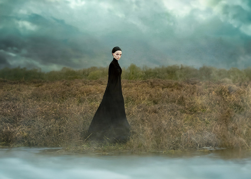 A witch in a long black gown is standing in a bog and staring menacingly at the viewer
