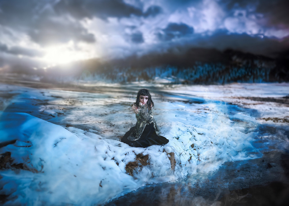 A young person with long dark hair and dramatic makeup sits on an icy, snow-covered ground with patches of water, under a dramatic dark sky with a bright sunburst. Their dark clothing blends with the