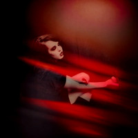 Portrait of a person with very pale skin and dark, dramatic eye and lip makeup, reclining in a dark room with strong red lighting and shadows, evoking a gothic or melancholic mood.