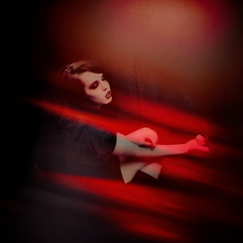 Portrait of a person with very pale skin and dark, dramatic eye and lip makeup, reclining in a dark room with strong red lighting and shadows, evoking a gothic or melancholic mood.