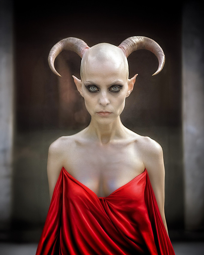 Aeshma, the female daemon of wrath, stands in eerie contrast—her pale, bald head adorned with downward-curving horns, a haunting visage of rage and chaos. Draped in a deep red silk dress, the fabric catches the light, flowing like spilled blood. Her presence is both mesmerizing and terrifying, embodying the seductive allure of destruction and the untamed fury of the unseen world.