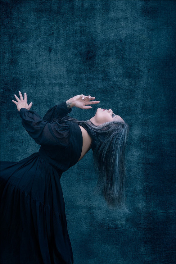 A person with long, dark, streaked hair and a flowing black dress is shown from the side, bending backward with arms raised, hands splayed, against a textured dark blue background. The pose suggests r