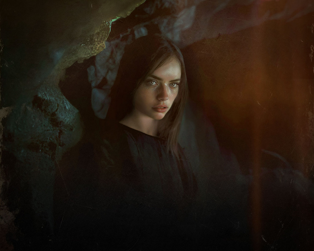 A young witch with long, straight dark brown hair gazes solemnly at the viewer, shrouded in a black robe. Behind her, a cavern’s rocky surface fades into shadows, while an eerie orange-red glow fills