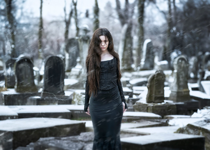 Pale witch with long brown hair in a fitted black gothic dress stands facing forward in a snowy, windy graveyard, surrounded by tombstones and barren trees, heavy snow falling at an angle.