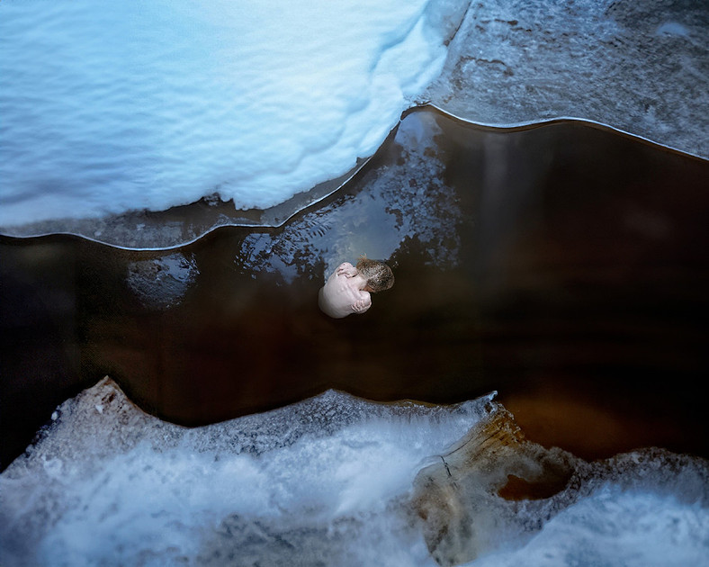 A blonde-haired witch sits in an icy river, her bare back exposed as she wraps her arms around herself in silent ritual. The frozen landscape stretches on either side, untouched snow framing the chill